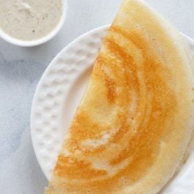 Ghee baked dosa folded and served on a white plate with a small bowl of coconut chutney with the text stay on it.