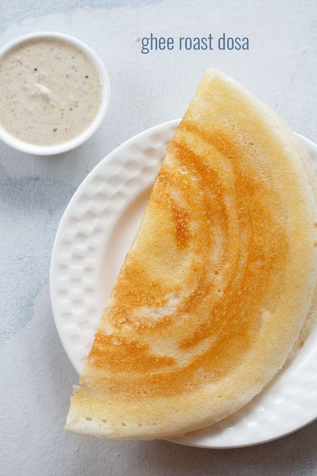 ghee roast dosa folded and served on a white plate with a small bowl of coconut chutney kept on the top left side with text layover.