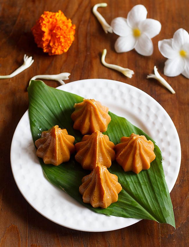 mawa modak served on a fresh turmeric leaf kept on a white plate with flowers in the background. 
