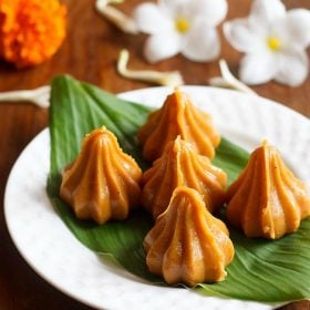 mawa modak served on a fresh turmeric leaf kept on a white plate with flowers in the background.