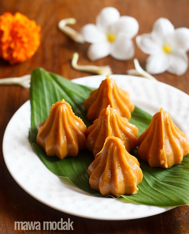 mawa modak served on a fresh turmeric leaf kept on a white plate with flowers in the background.