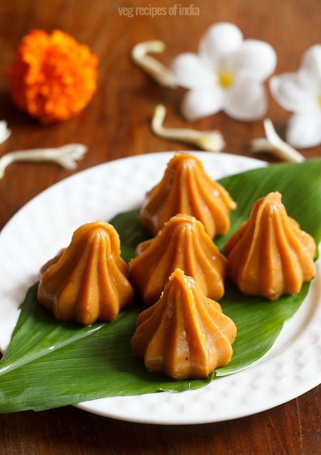 mawa modak served on a fresh turmeric leaf kept on a white plate with flowers in the background and text layovers. 
