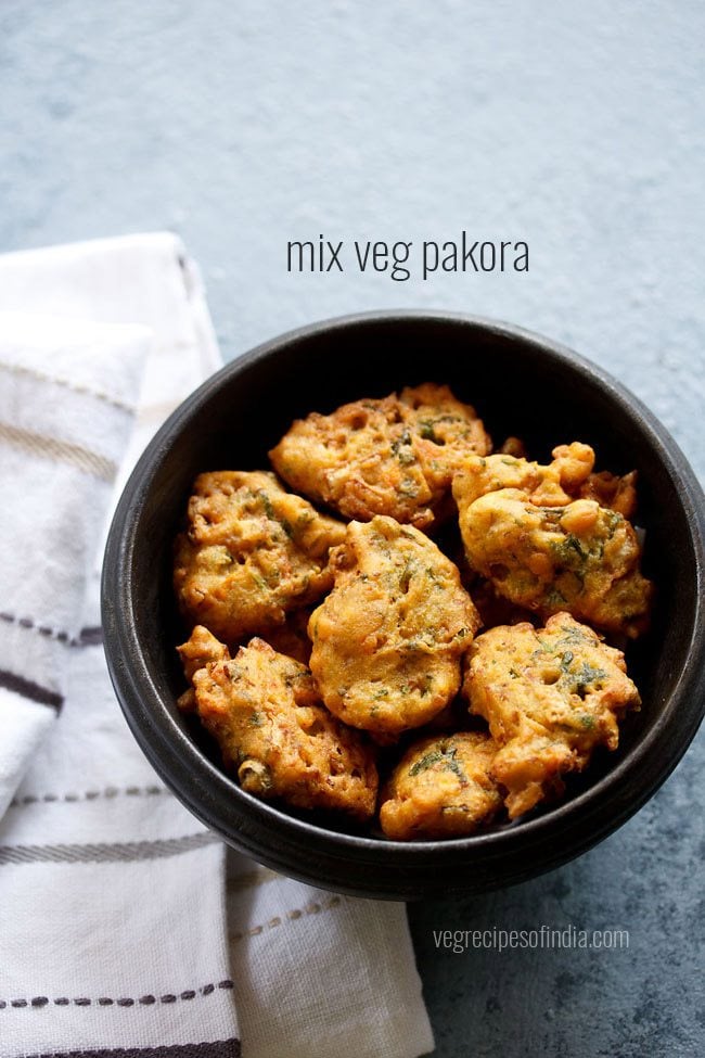 vegetable pakora in a black wooden bowl placed next to a white striped kitchen linen with text layover.