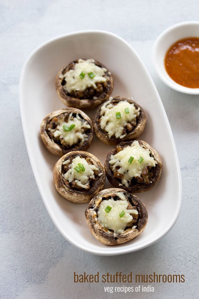 six baked stuffed mushrooms in an oblong serving bowl next to a ramekin of chutney. 