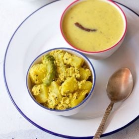 aloo posto served in a blue rimmed bowl on a white plate with a spoon kept on the right side and a bowl of dal kept on the top right side.