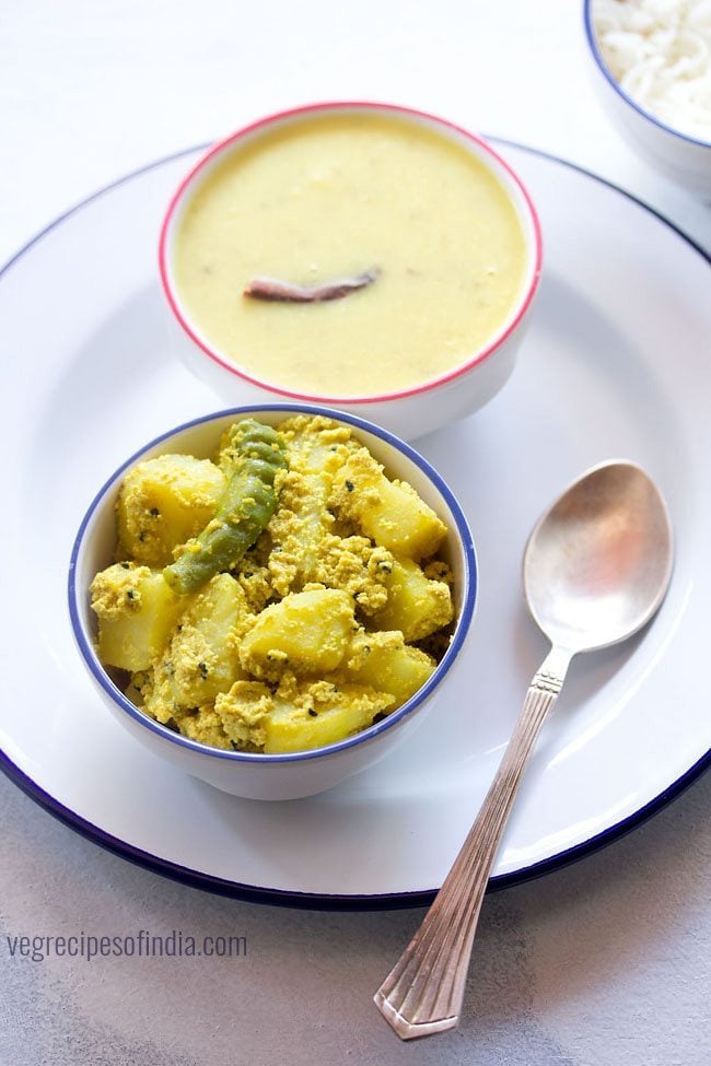 aloo posto served in a blue rimmed bowl on a white plate with a spoon kept on the right side, a bowl of dal kept on the top right side and text layover. 