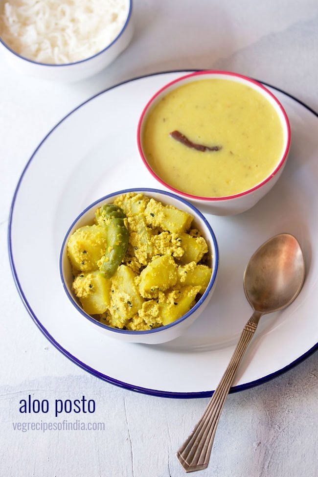 aloo posto served in a blue rimmed bowl on a white plate with a spoon kept on the right side and a bowl of dal kept on the top right side.