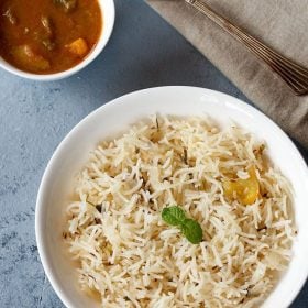 kuska rice garnished with a mint sprig and served in a white plate with a bowl of curry on the top left side.