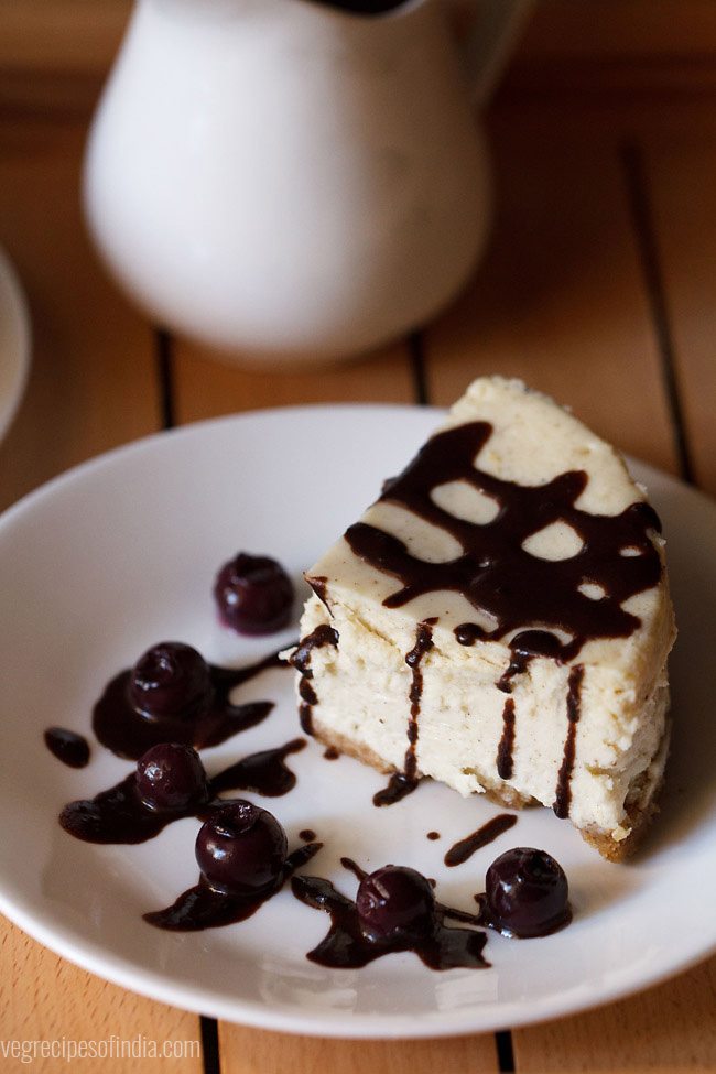 wedge of New York Style cheesecake on a serving plate that has been drizzled with homemade chocolate sauce and garnished with blue berries.
