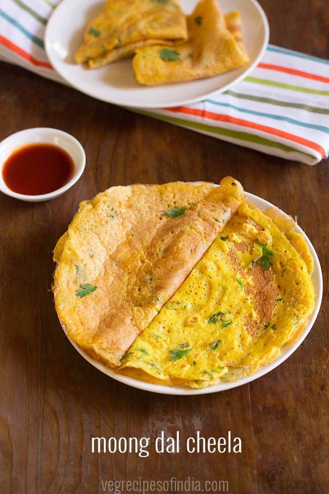 moong dal chilla folded into halves and garnished with coriander leaves, served on white plate with a small bowl of tomato ketchup kept on the top left side and text layovers. 