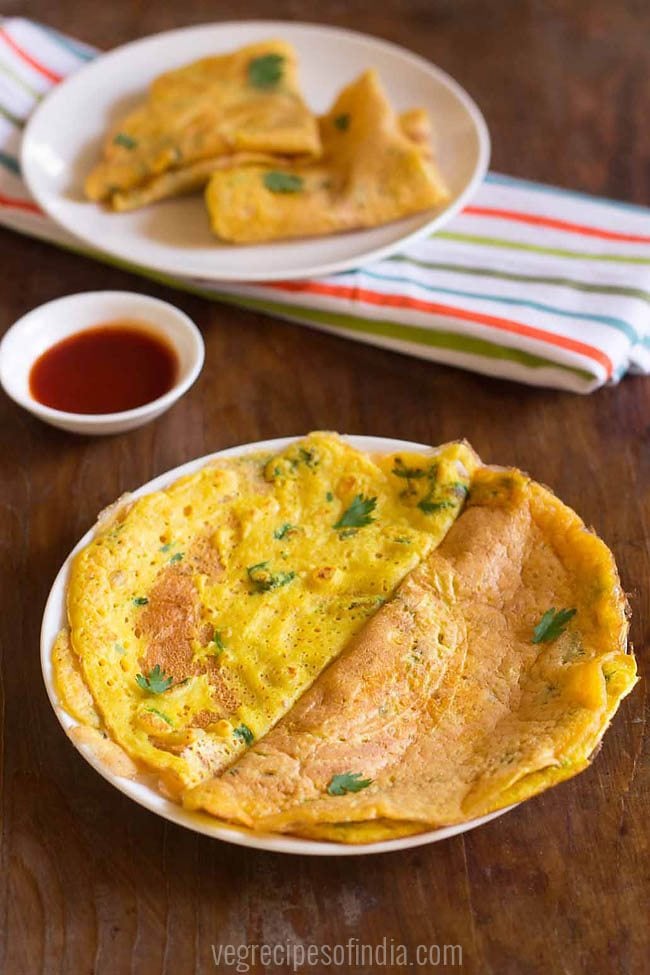 moong dal chilla folded into halves and garnished with coriander leaves, served on white plate with a small bowl of tomato ketchup kept on the top left side and text layover. 