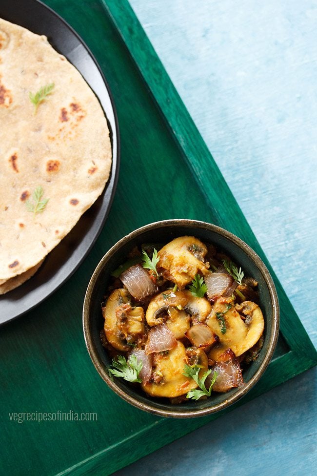 mushroom do pyaza garnished with cilantro sprigs in a dark olive green ceramic bowl on a dark green board with a side of roti 