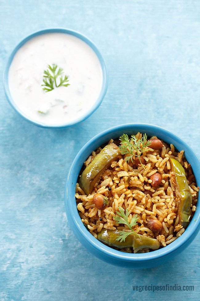 karnataka style vangi bath garnished with coriander leaves and served in a blue bowl with a light blue colored bowl of raita and text layover. 