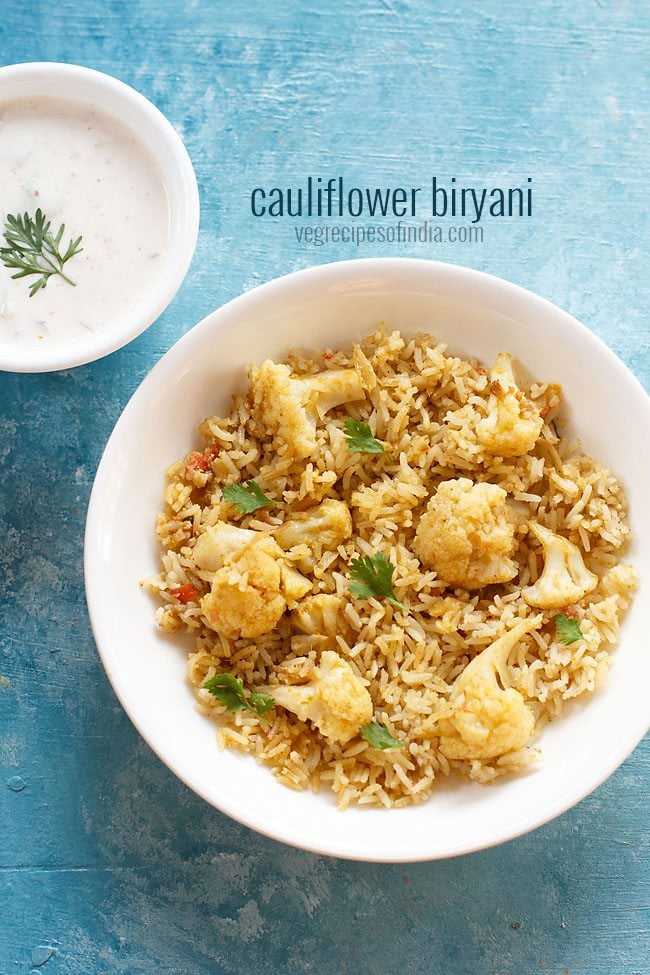 cauliflower biryani garnished with coriander leaves and served in a white bowl with a bowl of raita kept in the top left side and text layovers.