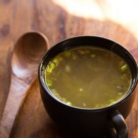 veg clear soup in a black bowl with a wooden soup spoon in the bowl on a wooden board.