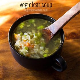 vegetable soup in a black bowl with a wooden soup spoon with some veggies on it in the bowl on a wooden board