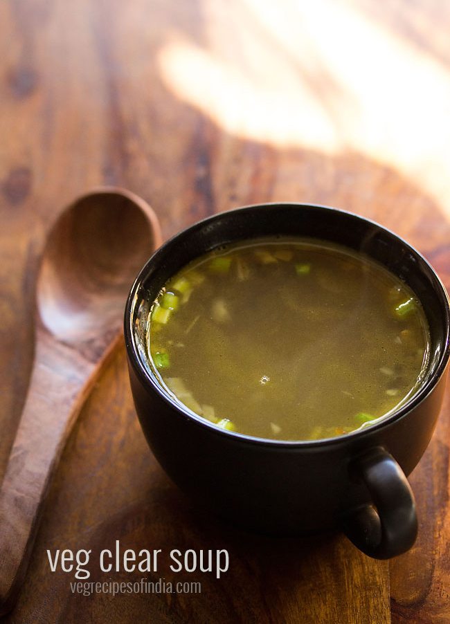 veg clear soup in a black bowl with a wooden soup spoon in the bowl on a wooden board.