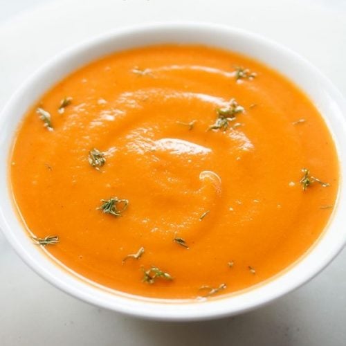 white bowl filled with carrot ginger soup and garnished with fresh green herbs.