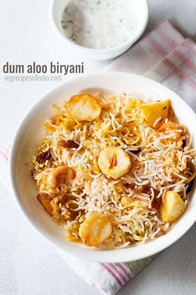 aloo biryani served in a white plate. with a bowl of raita kept on the top left side and text layovers.