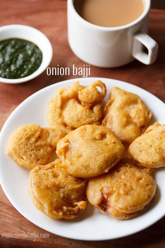 onion bajji served on a white plate with a bowl of green chutney and a cup of tea kept in the background and text layover.