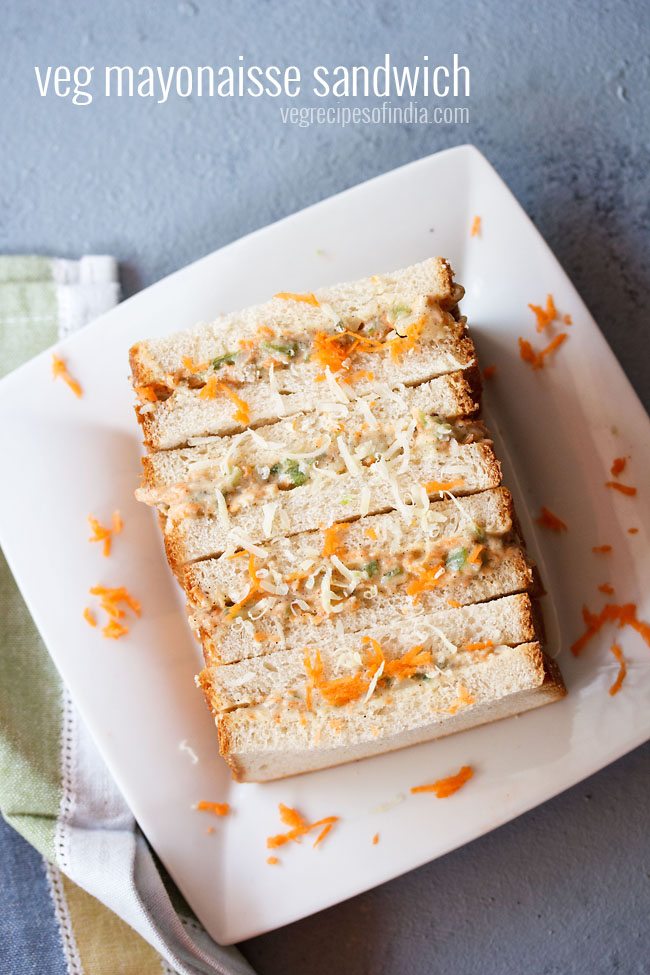 veg mayonnaise sandwich topped with grated carrots and cheese on a square white plate