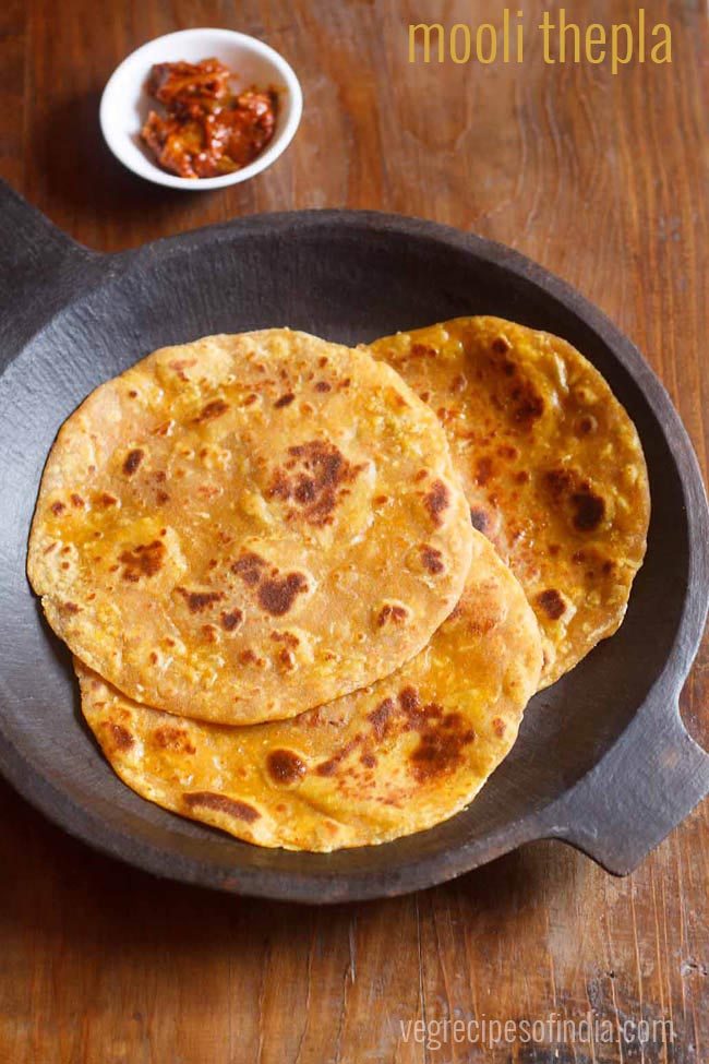 mooli thepla served in a black round stone tray with a side of mango pickle in a small white bowl on a brown wooden board and text layover. 