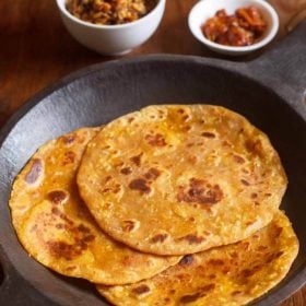 mooli thepla placed in an black round stone tray with a side of mango pickle in a small white bowl, dry chutney in another white bowl on a brown wooden board