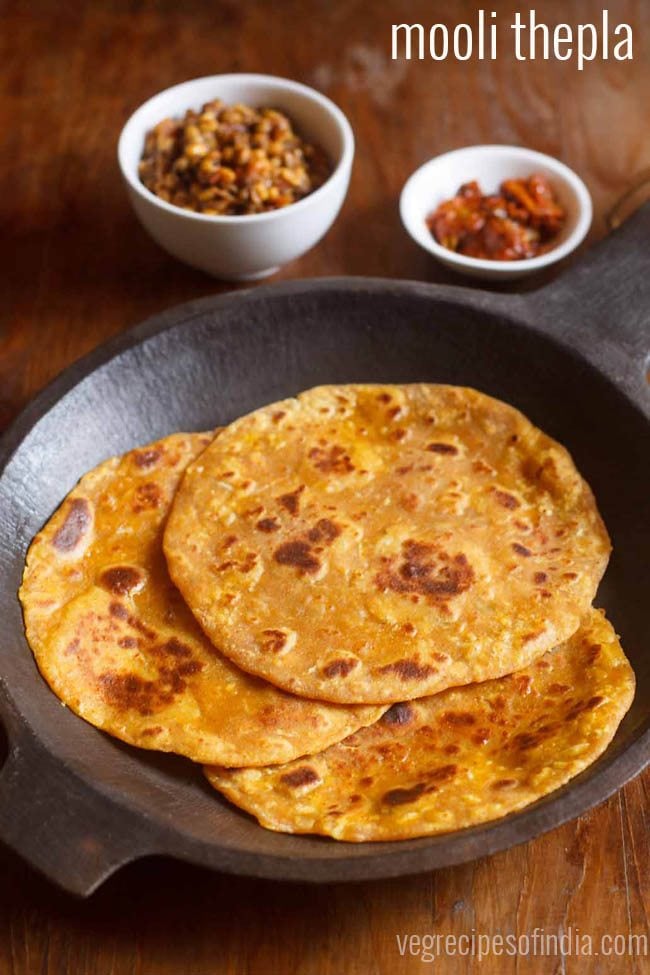 mooli thepla placed in an black round stone tray with a side of mango pickle in a small white bowl, dry chutney in another white bowl on a brown wooden board