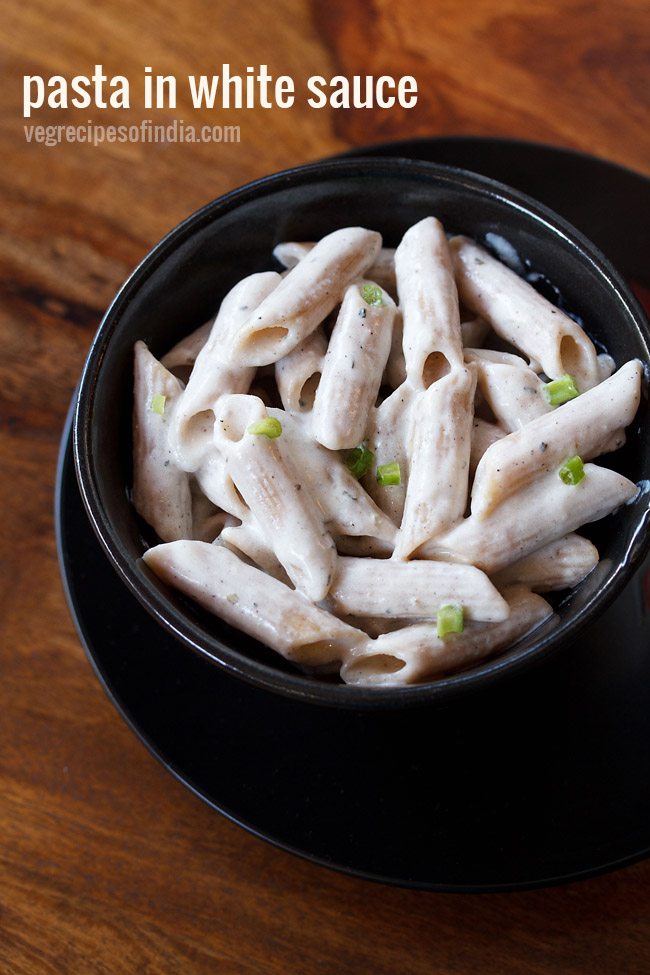 white sauce pasta garnished with scallion greens served in a black glass bowl kept on top of black plate on a wooden board with text layovers. 