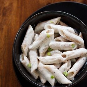 white sauce pasta garnished with scallion greens served in a black glass bowl kept on top of black plate on a wooden board