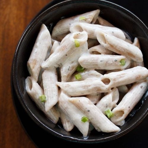 white sauce pasta garnished with scallion greens served in a black glass bowl kept on top of black plate on a wooden board