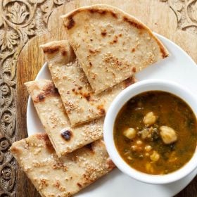 cheese garlic naan cut into wedges and served on a white plate with a bowl of curry kept on the right side.