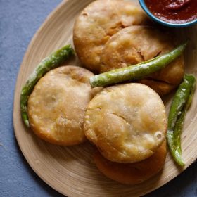 dal kachori placed on a wooden platter with fried green chilies and red chili chutney in a bowl.