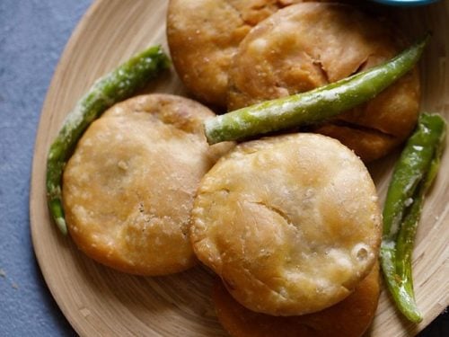 dal kachori placed on a wooden platter with fried green chilies and red chili chutney in a bowl.