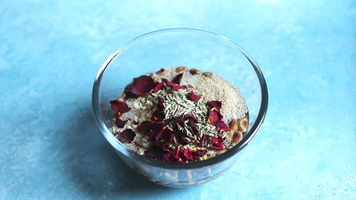 fennel seeds on top of rose petals floating in a glass bowl placed on a bright light blue board