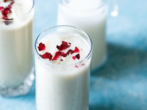 thandai recipe garnished with rose petals in two glass jars with a small glass jar of milk in the background on a bright blue board