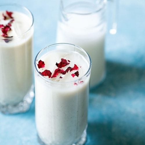 thandai recipe garnished with rose petals in two glass jars with a small glass jar of milk in the background on a bright blue board