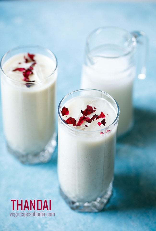 thandai garnished with rose petals served in 2 glasses with a small glass jar of milk in the background on a bright blue board and text layover. 