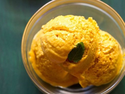 close up shot of scoops of mango ice cream in a glass bowl garnished with a mint leaf on a green board