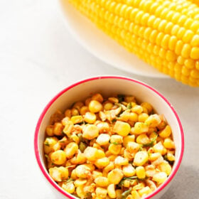 Corn salsa served in a white bowl with dark pink rim on white background with steamed corn on the cob next to it