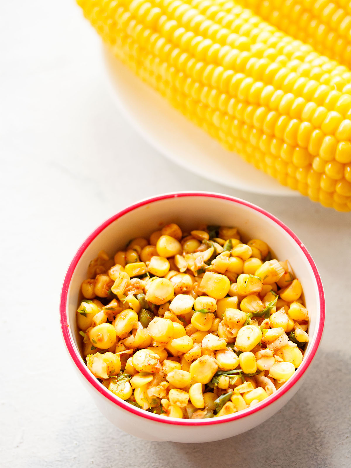 corn salsa served in a dark pink rimmed white bowl on a white background with a steamed corn cob at the side