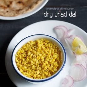 sukhi urad dal served in a blue rimmed white bowl placed on a white plate, onion slices and lemon wedges kept on right side on the plate, parathas and text layovers.