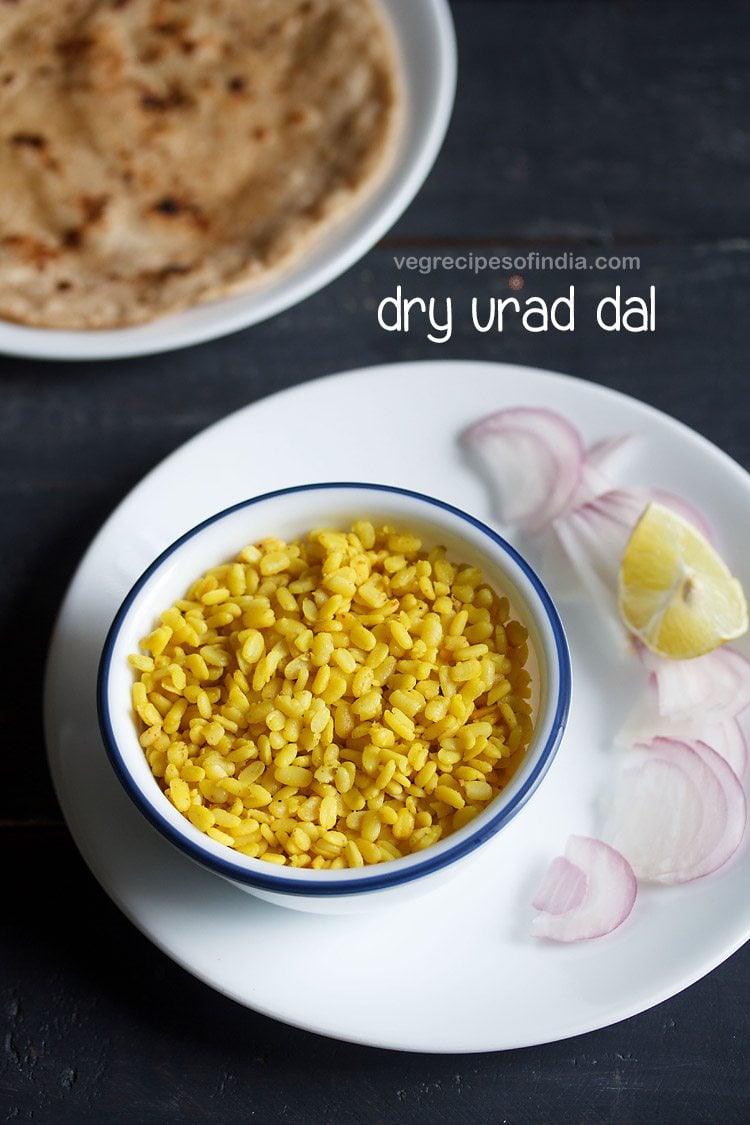 urad dal recipe served in a blue rimmed white bowl placed on a white plate, onion slices and lemon wedges kept on right side on the plate, parathas and text layovers.