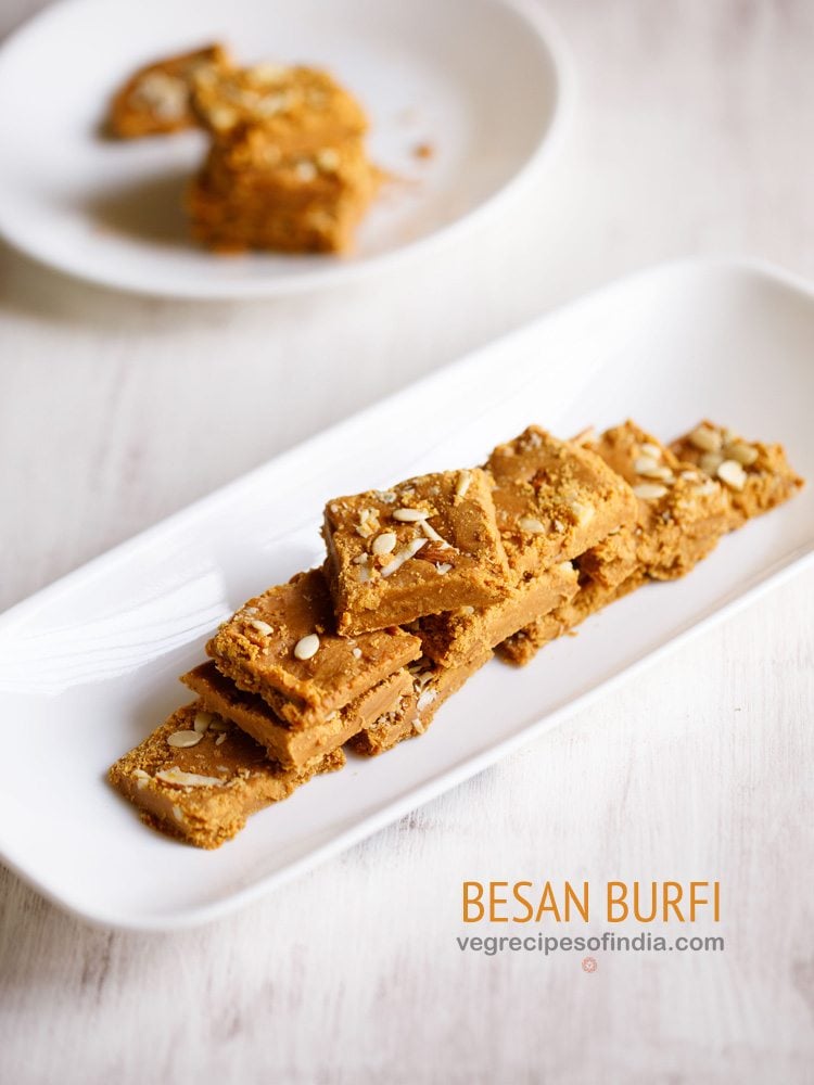 besan ki barfi pieces stacked on top of one another and served on a rectangular white platter with text layovers.