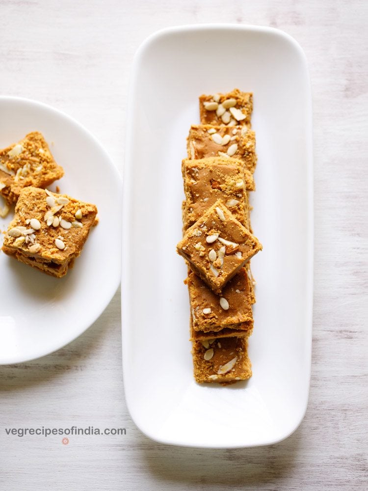 top shot of besan ki barfi pieces stacked on top of one another and served on a rectangular white platter with text layover. 