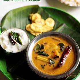 varutharacha sambar served in a copper bowl placed on a plate lined with banana leaf and bowl of pachadi, side of chips and text layovers.