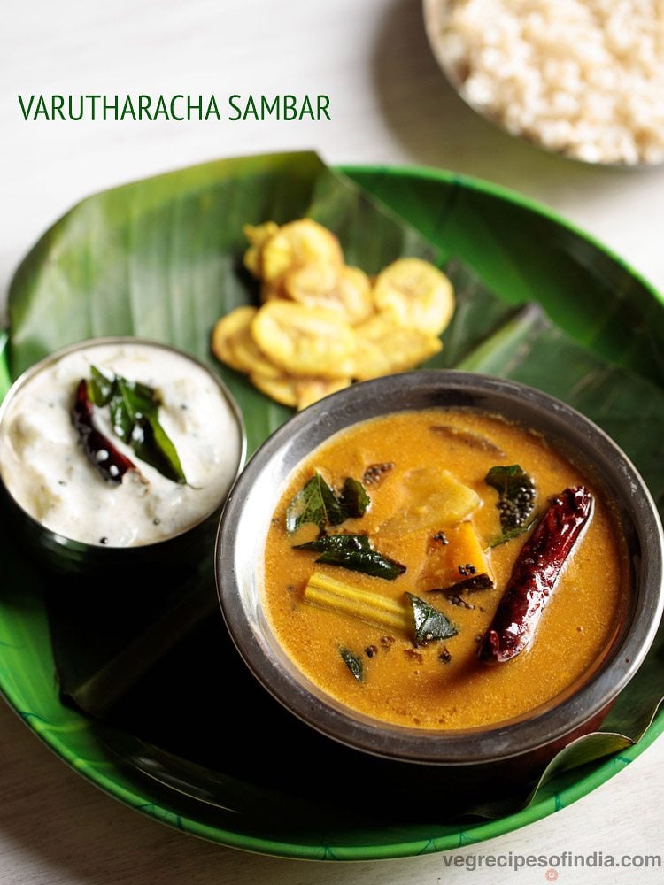 varutharacha sambar served in a copper bowl placed on a plate lined with banana leaf and bowl of pachadi, side of chips and text layovers.