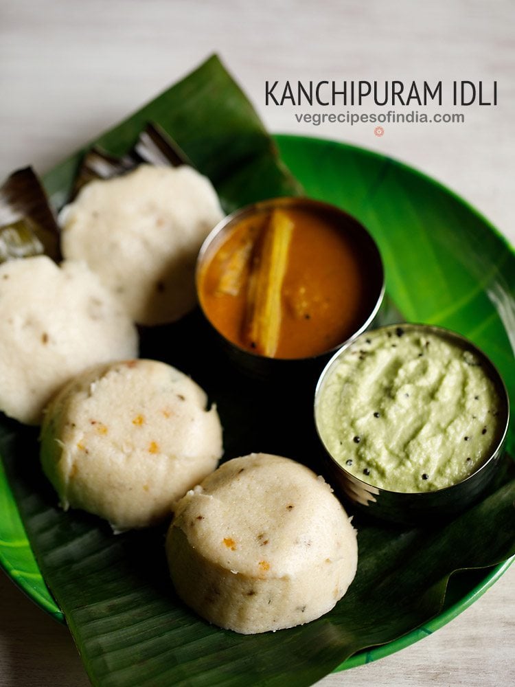 kanchipuram idli served on a banana leaf placed on a green colored plate with a bowl of coconut chutney and a bowl of sambar kept on the right side on the plate and text layovers.
