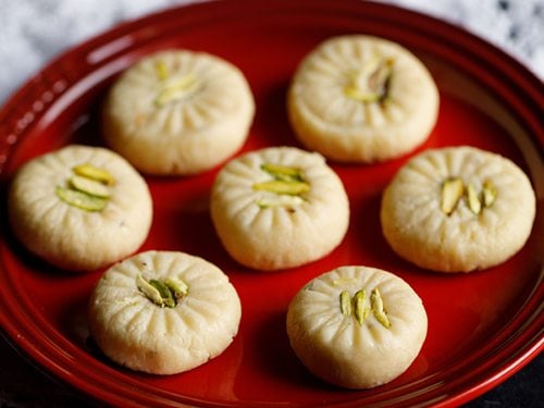 closeup shot of 7 milk peda pieces on a red plate placed on white doilies
