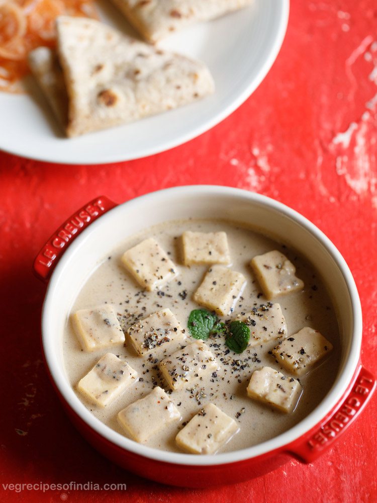 paneer kali mirch garnished with mint leaves and crushed black pepper and served in a red ceramic casserole with a plate of chapattis kept on the top left side and text layover. 